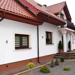 Biały dom z brązowym cokołem, czerwonym dachem i ozdobnymi schodami.