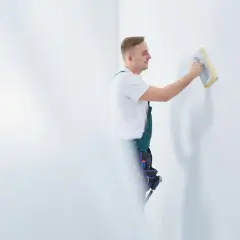 Fachowiec w zielonym ogrodniczkach wykonuje szlifowanie gładzi na białej ścianie, przygotowując ją do malowania.