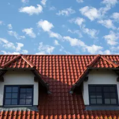 Dachówka ceramiczna w kolorze ceglastym pokrywa dach domu z dwoma lukarnami. Różne rodzaje dachówek tworzą harmonijną całość.