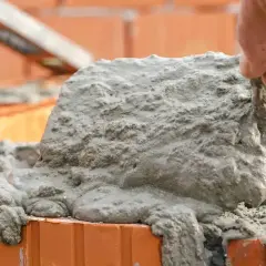 Ręka murarza nakłada świeżą zaprawę na cegły. Budowa trwa, a zaprawa spływa po bokach.