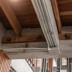 Metalowe rury elektryczne biegną wzdłuż betonowego podciągu, łącząc się w puszce. Drewniane belki stropowe i beton tworzą surową, industrialną estetykę.