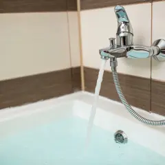 Chromowana bateria wannowa z wylewką i słuchawką prysznicową, z której leje się woda do wanny. Widać płytki na ścianie i wannę. Wysokość baterii wannowej jest standardowa.