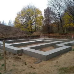 Fundament domu wylewany na ławach fundamentowych, poniżej strefa przemarzania gruntu. W tle jesienne drzewa i usypana ziemia.