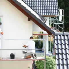 Dwóch mężczyzn na rusztowaniu maluje białą elewację domu. Sprawdź, jaka jest malowanie elewacji cena.