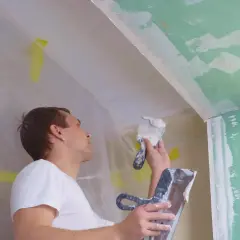 Mężczyzna z **pacą** wygładza tynk na suficie. Wokół folia ochronna.