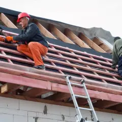 Dwaj budowlańcy pracują na dachu, montując wiatroizolację. Jeden z nich w pomarańczowych spodniach i czerwonym kasku wbija gwoździe.