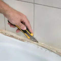 Ręka z nożem usuwa stary silikon z wanny. Dowiedz się, czym usunąć silikon, by odnowić łazienkę.