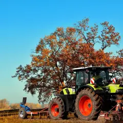 Traktor z pługiem na polu, przygotowujący teren pod przekształcenie działki rolnej na budowlaną. Jesienne drzewo w tle.