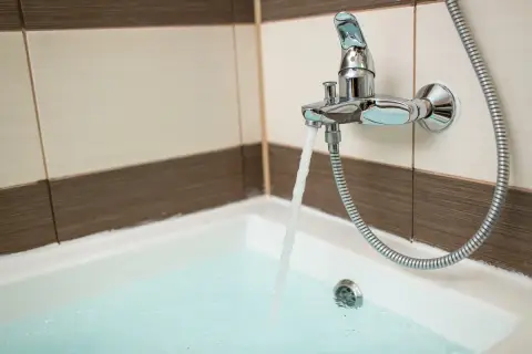 Chromowana bateria wannowa z wylewką i słuchawką prysznicową, z której leje się woda do wanny. Widać płytki na ścianie i wannę. Wysokość baterii wannowej jest standardowa.