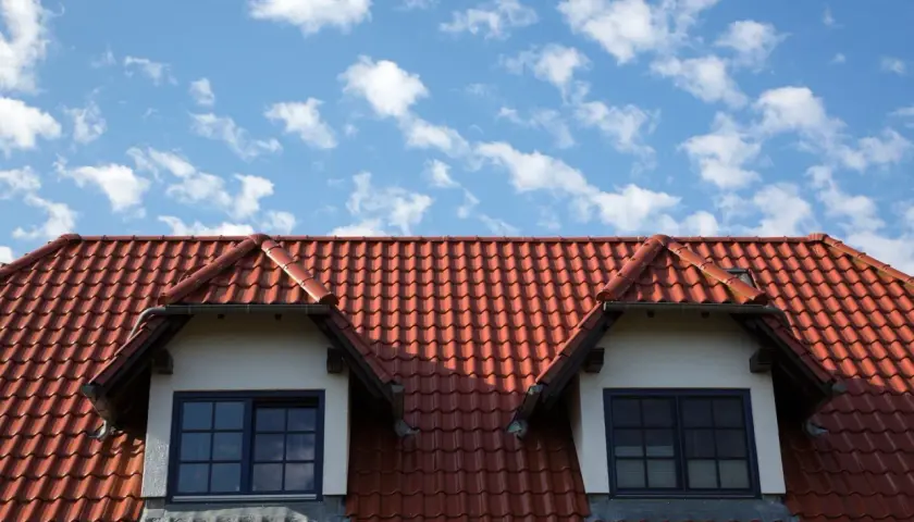 Dachówka ceramiczna w kolorze ceglastym pokrywa dach domu z dwoma lukarnami. Różne rodzaje dachówek tworzą harmonijną całość.