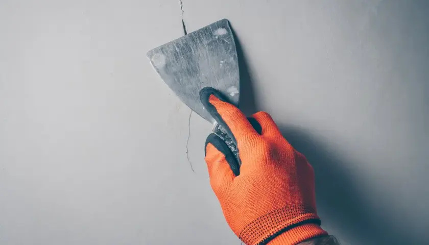 Ręka w pomarańczowej rękawicy wypełnia głębokie pęknięcia w ścianie szpachlą za pomocą kielni.