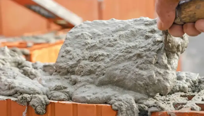 Ręka murarza nakłada świeżą zaprawę na cegły. Budowa trwa, a zaprawa spływa po bokach.