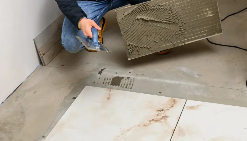 Układanie płytek 60x60 cennik. Majster z kielnią nakłada klej na płytkę, przygotowując ją do położenia na podłodze.