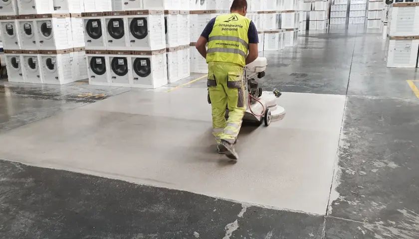 Pracownik w żółtym odblaskowym stroju szlifuje betonową posadzkę w magazynie. Sprawdź szlifowanie betonu cena za m2.