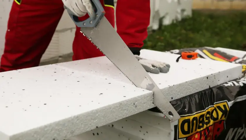 Ręce w rękawiczkach tną styropian piłą ręczną. W tle widać miarkę i materiały budowlane.