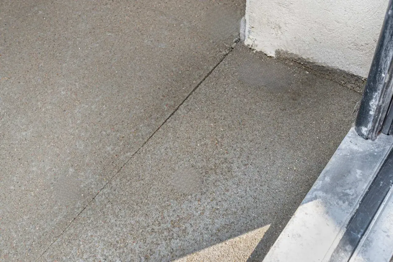 Szara, ziarnista powierzchnia, prawdopodobnie beton, z widocznym pęknięciem. Obok biała ściana i fragment czarnej poręczy. Mastyka użyta do uszczelnienia.