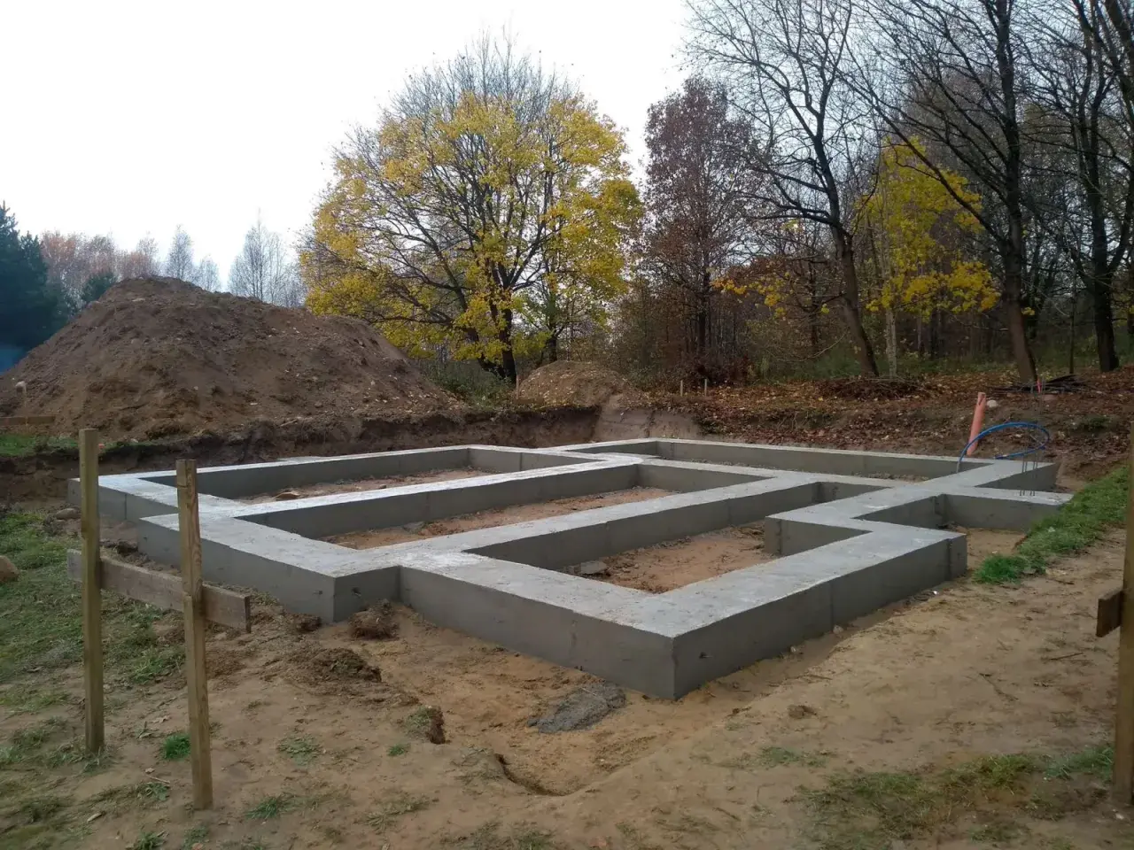 Fundament domu wylewany na ławach fundamentowych, poniżej strefa przemarzania gruntu. W tle jesienne drzewa i usypana ziemia.