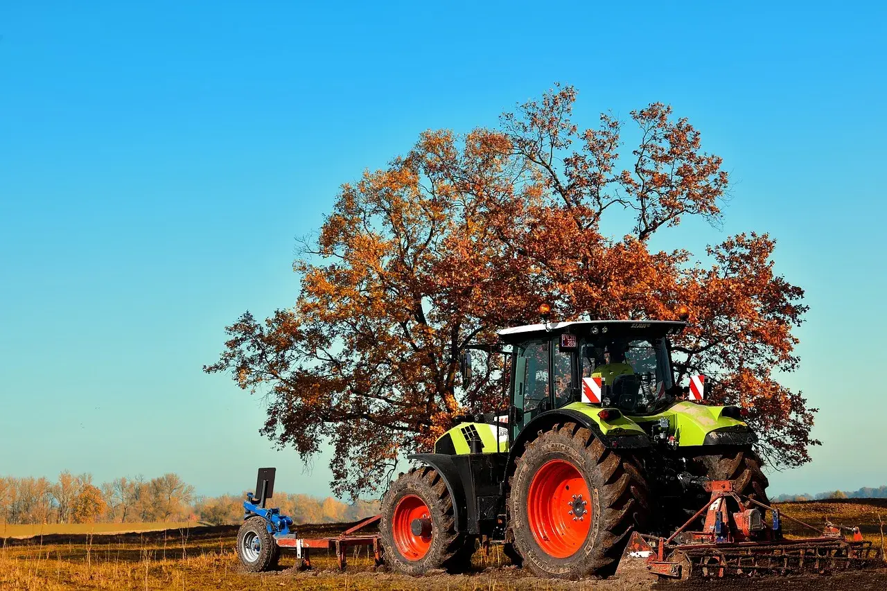 Traktor z pługiem na polu, przygotowujący teren pod przekształcenie działki rolnej na budowlaną. Jesienne drzewo w tle.
