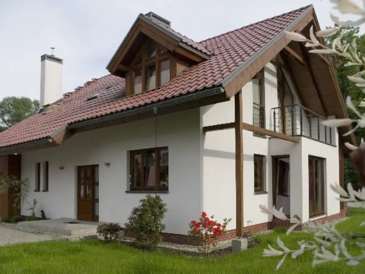 Nowoczesny dom z białym tynkiem i drewnianymi elementami. Widoczna jest ciekawa struktura elewacyjna, dachówka i balkon.