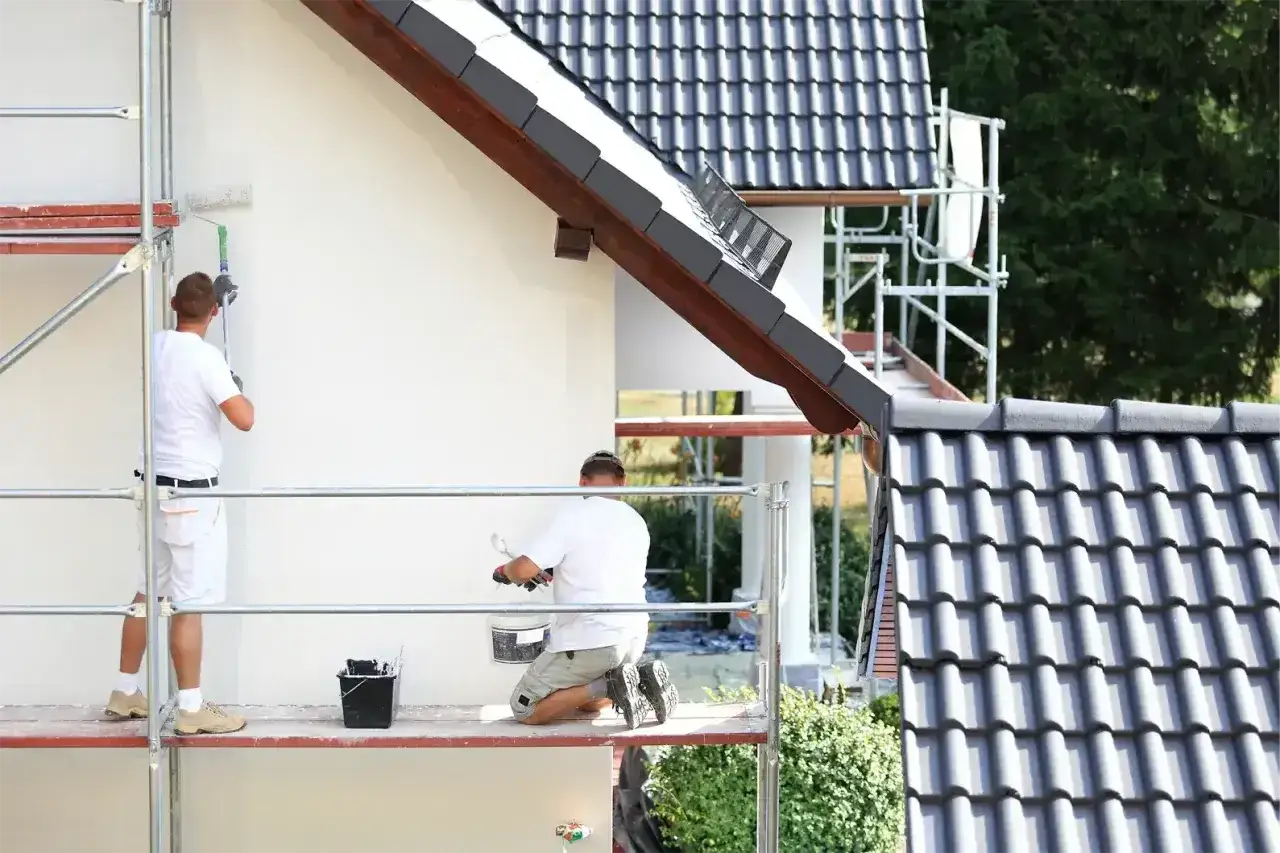 Dwóch mężczyzn na rusztowaniu maluje białą elewację domu. Sprawdź, jaka jest malowanie elewacji cena.