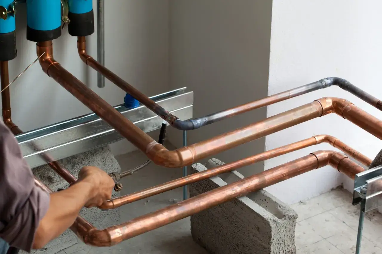 Instalator lutuje miedziane rury. Widać fragmenty instalacji, niebieskie filtry i betonowe podpory.